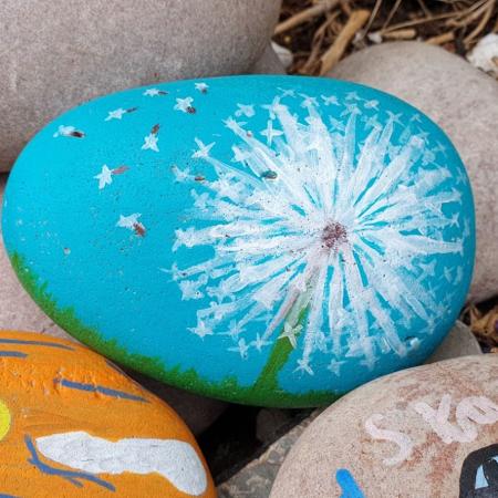 pebbles, the top one is painted to show dandelion seeds blwing away against a blue sky
